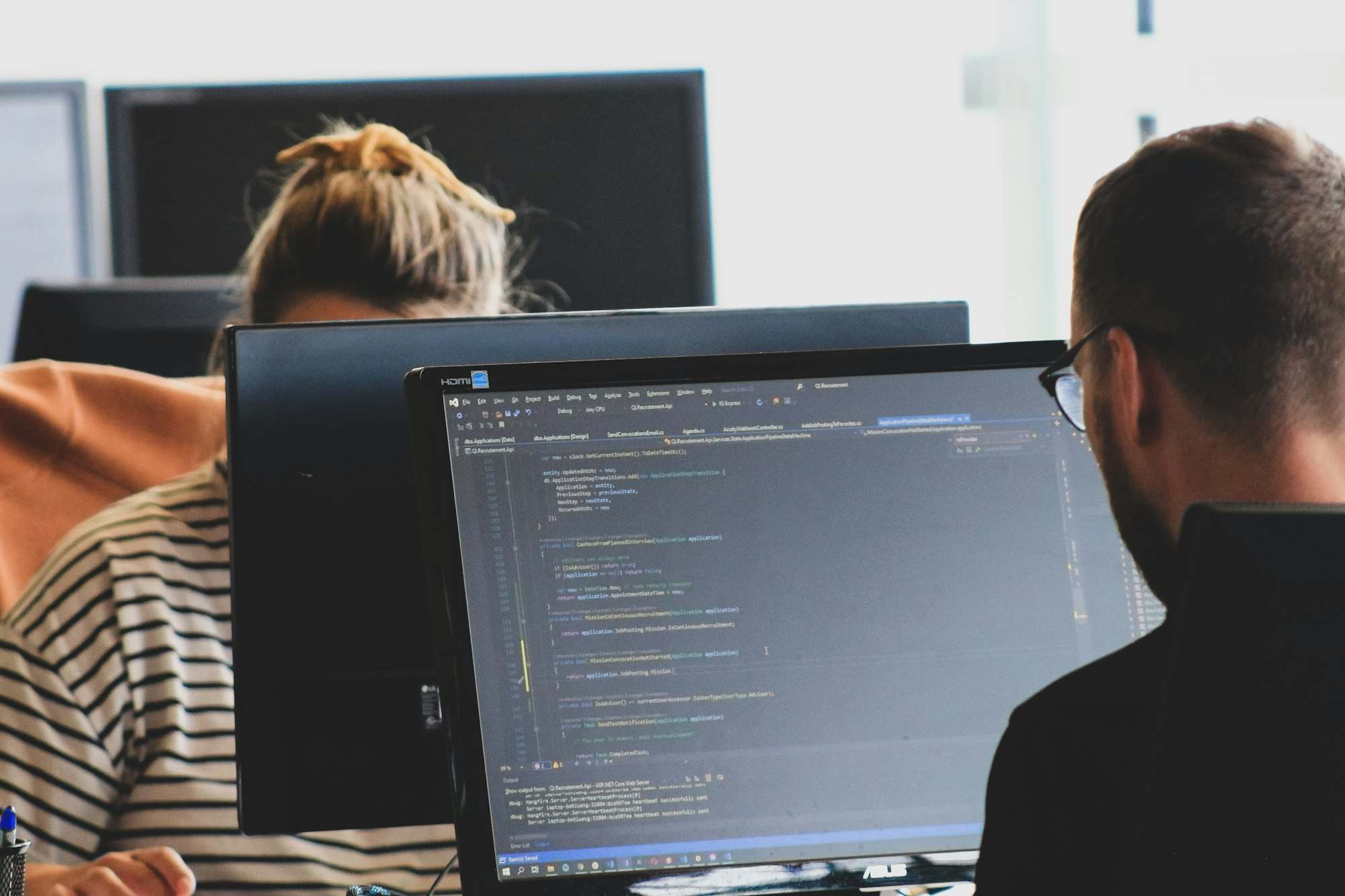 Zwei Personen arbeiten in einem hellen Büro an Monitoren mit Quelltext